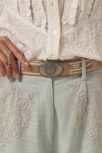 Close-up of a person wearing a beige belt with a large buckle, white lace shirt, and light-colored pants with embroidery.