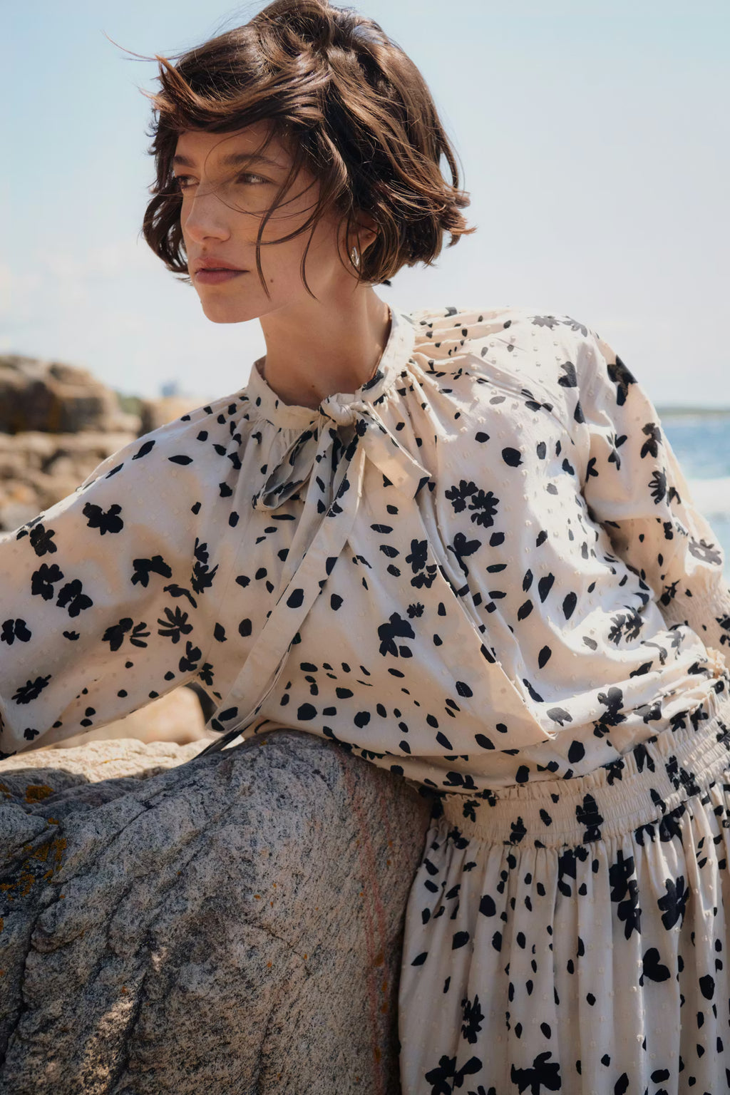 Woman wearing a floral dress sitting on a rock by the sea.