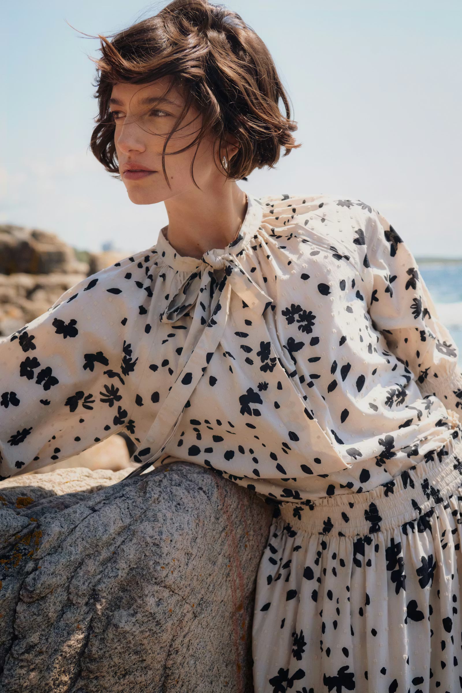 Woman wearing a floral dress sitting on a rock by the sea.