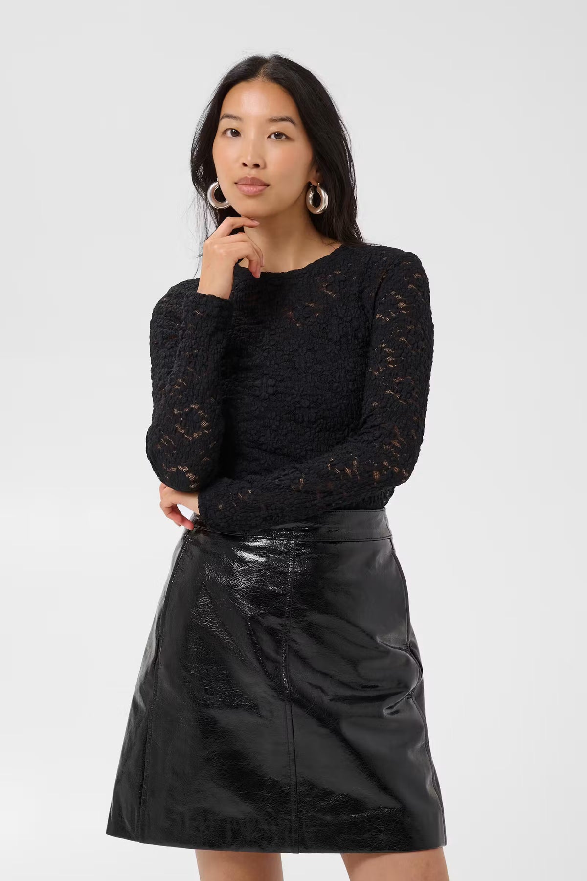 A woman modeling a black long-sleeve lace top paired with a black faux-leather mini skirt, set against a plain light gray studio background.