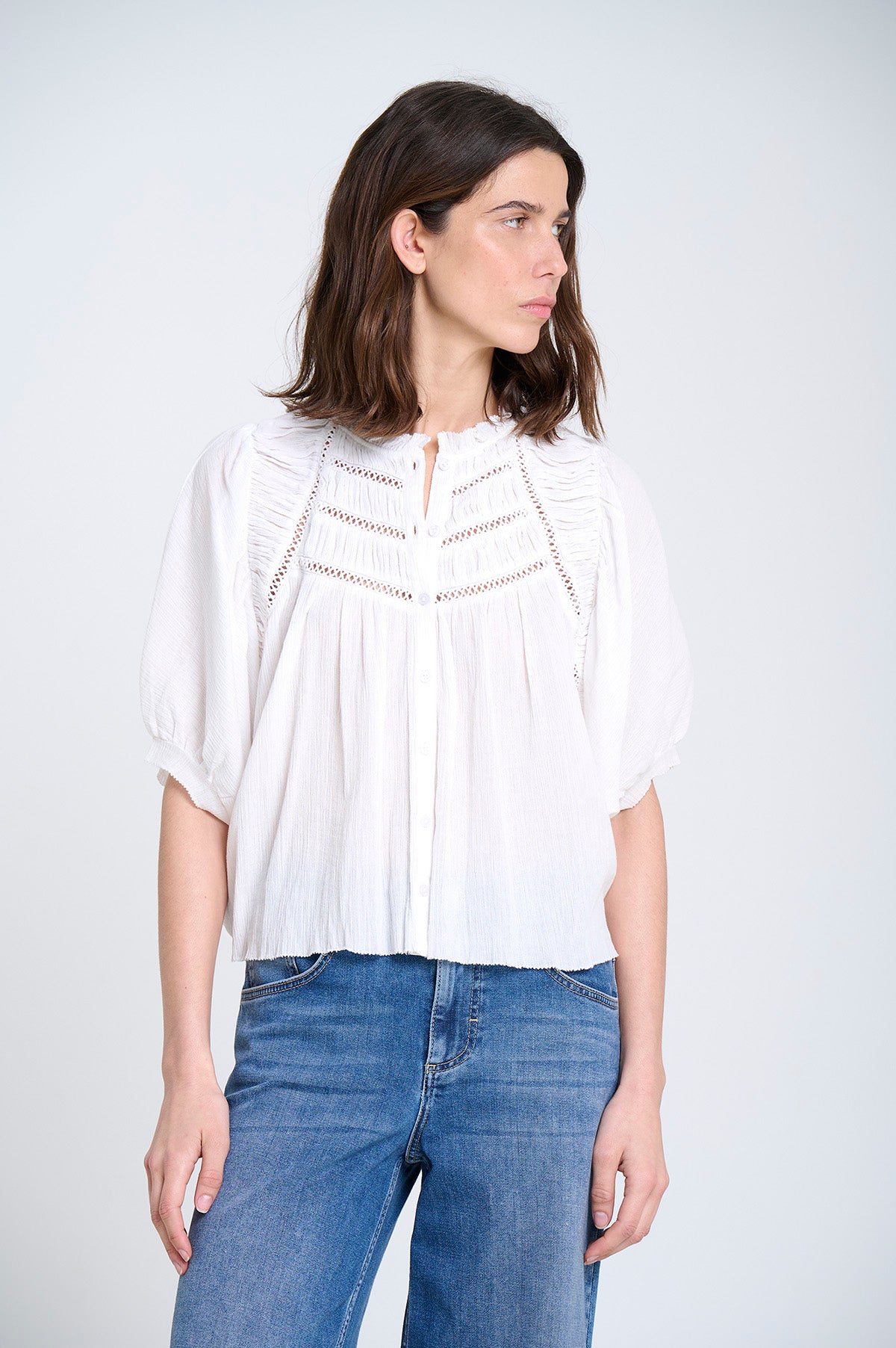 Woman wearing a white blouse with lace boho details and blue jeans on a plain background