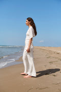 Woman in a white outfit standing on a beach with clear blue sky and ocean.