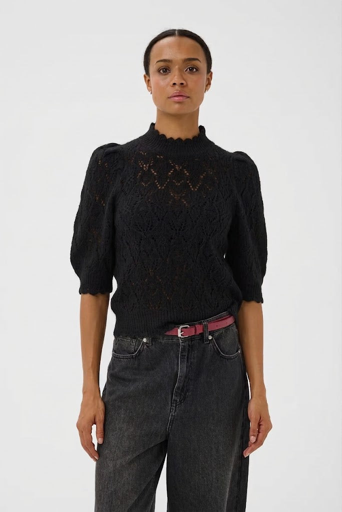 A model wearing a black lace knit mock-neck top with puffed 3/4 sleeves, paired with dark gray jeans and a pink belt, against a plain light gray background.