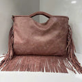 A dusty rose faux-leather fringe handbag with a top handle, displayed on a white surface against a neutral gray background.