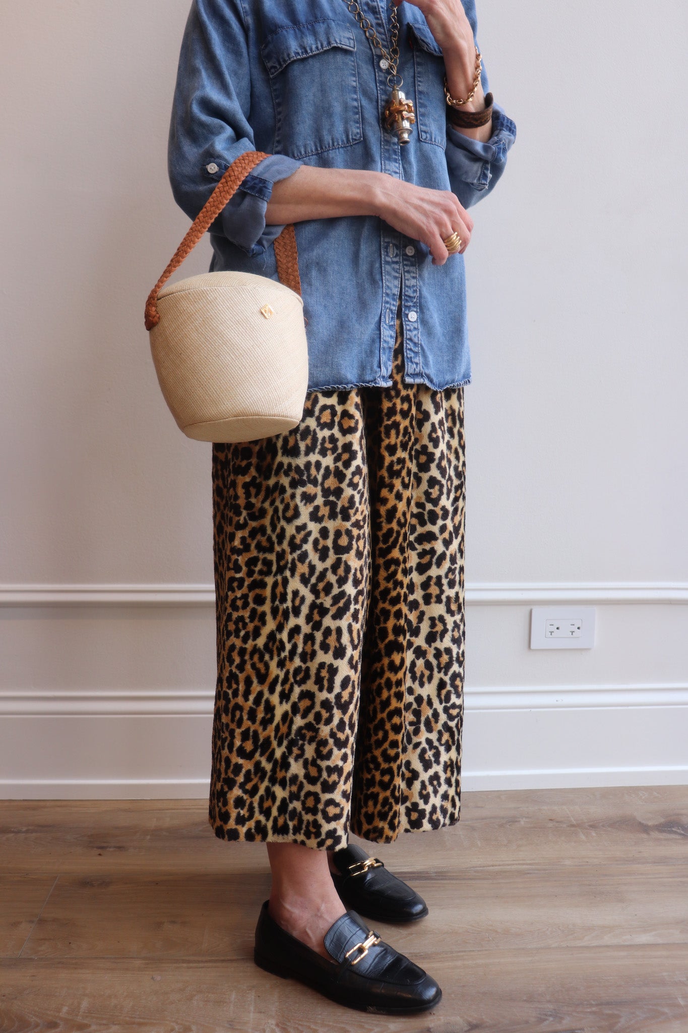 Person wearing leopard print pants and a denim jacket, holding a beige bucket handbag indoors.