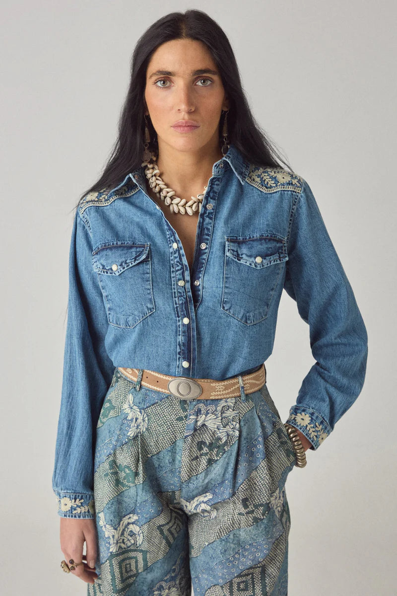 Woman wearing a blue denim shirt with floral patterns and a patterned skirt on a gray background