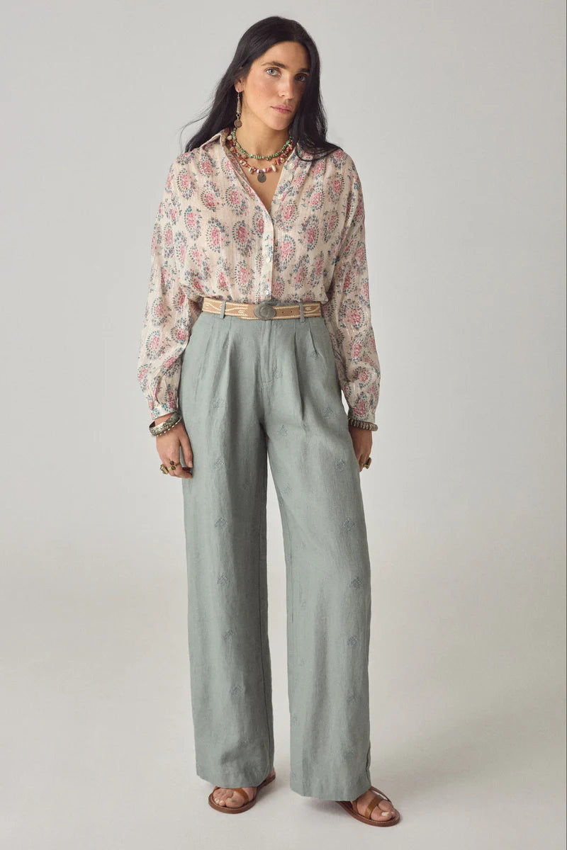 Woman wearing a patterned blouse and light gray pants on a plain background