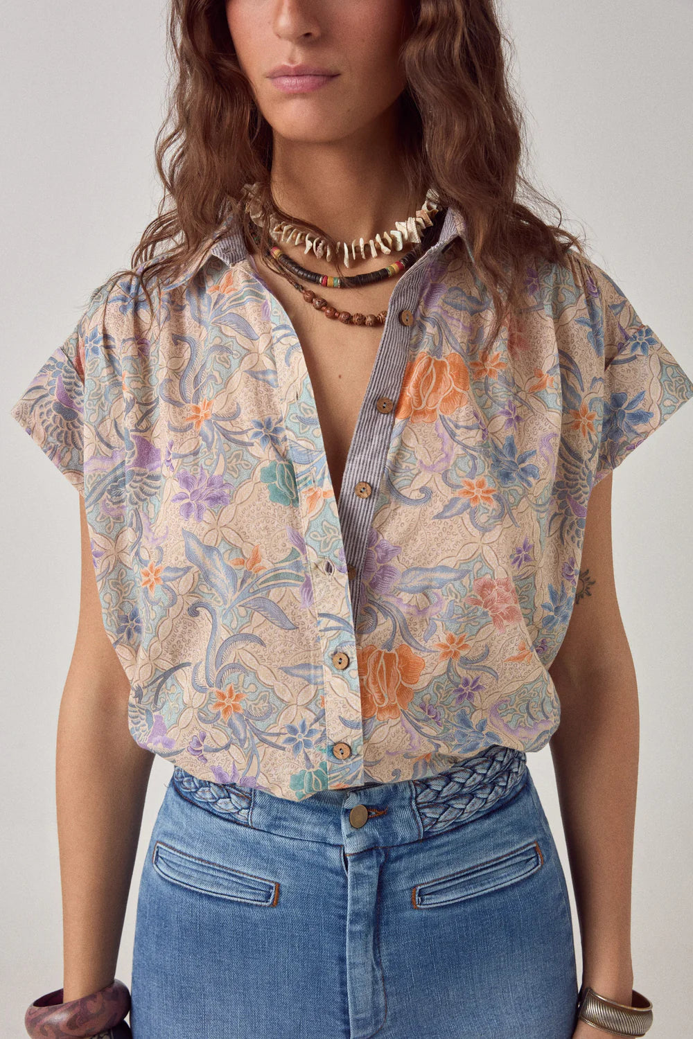 Woman wearing a patterned shirt and blue jeans against a plain background. Tropical Top, tropical floral button up cap sleeve top. 