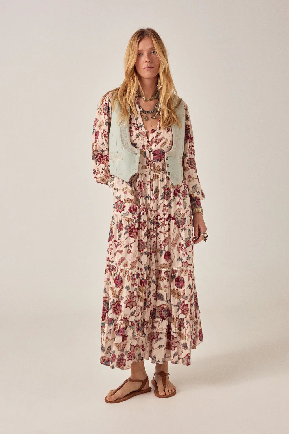 Woman wearing a floral dress on a plain background. menswear vest, blue vest, layering over a dress, dress layering, how to style a maxi dress 