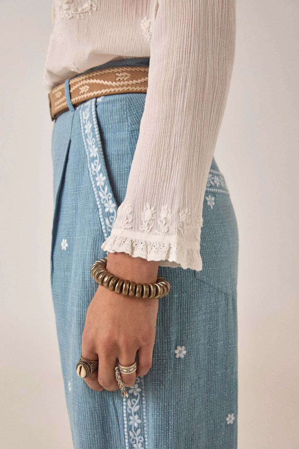 Person wearing a white long-sleeve top and blue denim skirt with floral embroidery, holding a gold bracelet and ring.
