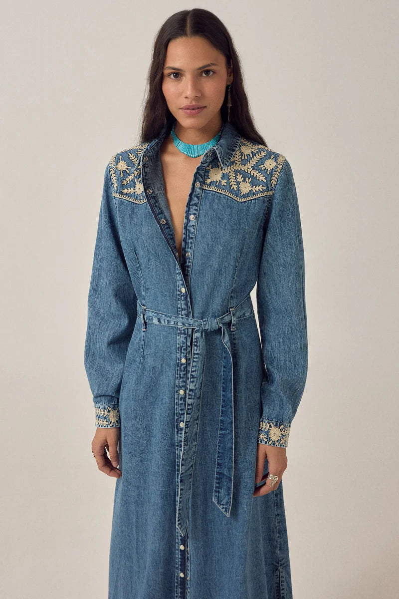 Woman wearing a long denim dress with floral embroidery on a plain background