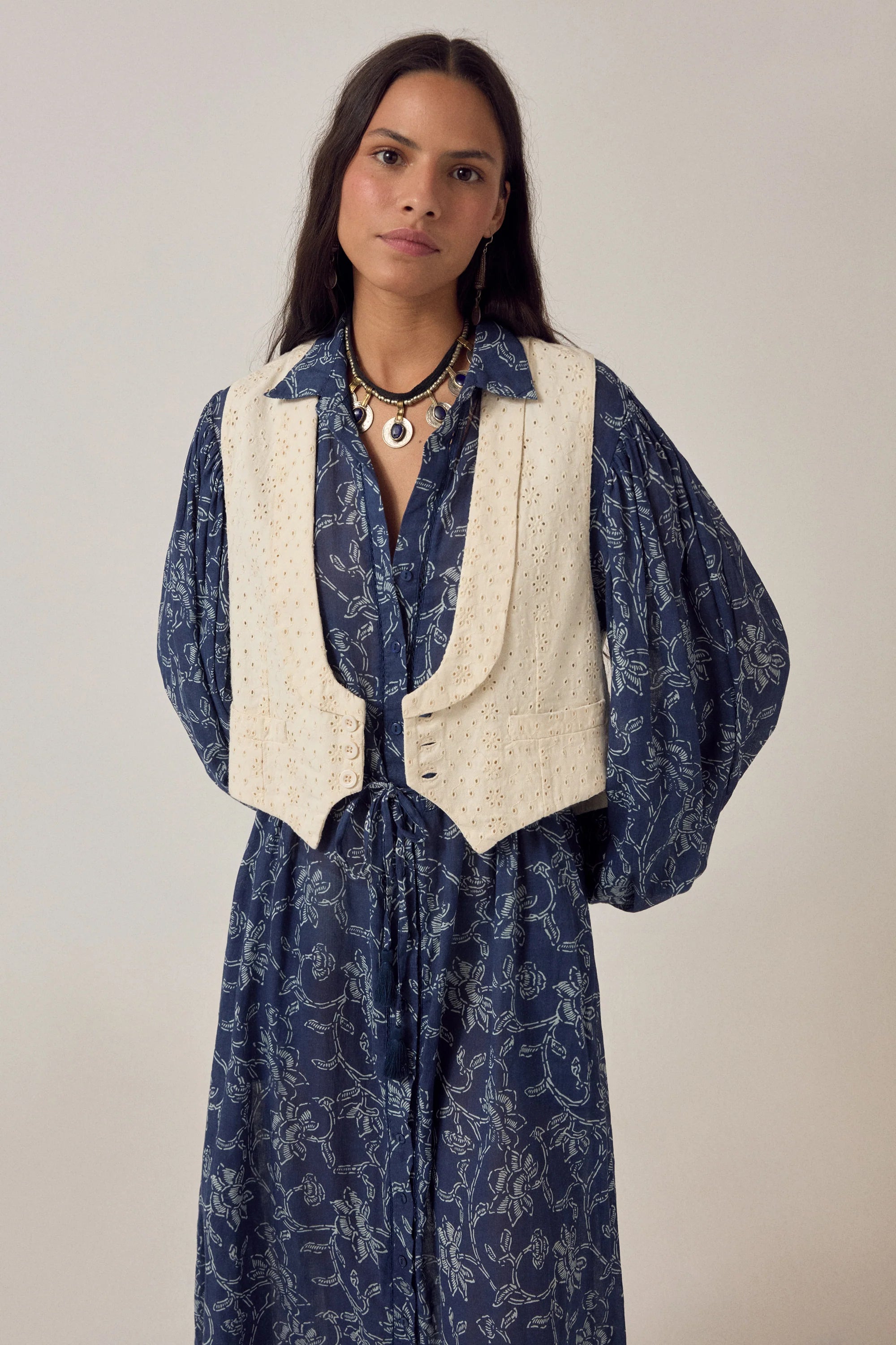 Woman wearing a blue patterned dress with a white vest against a plain background