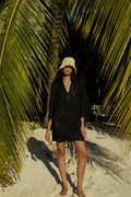 Woman in a black dress and white hat standing under palm trees on a sandy path.