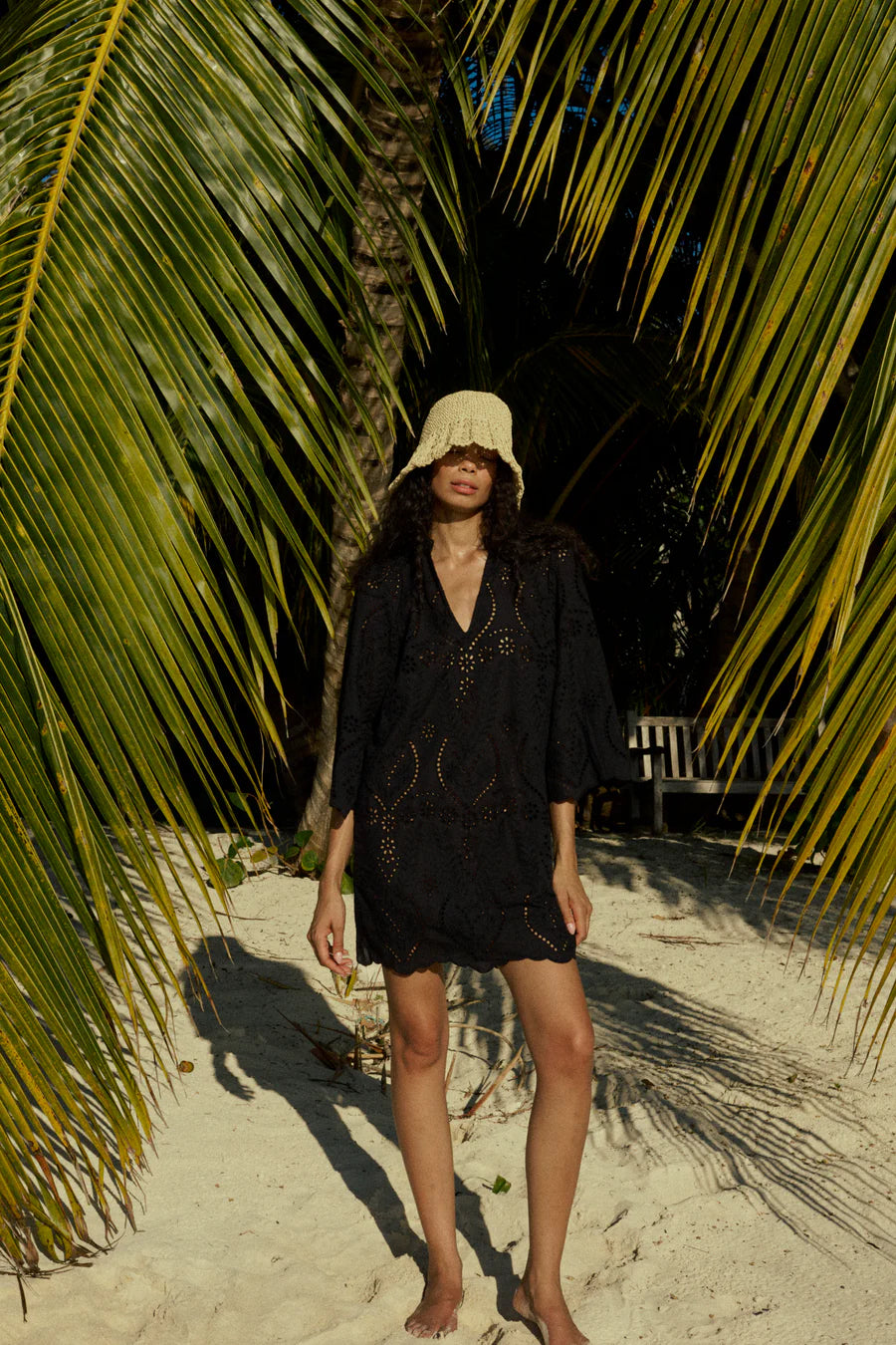 Woman in a black dress and white hat standing under palm trees on a sandy path.