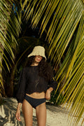 Woman in a black outfit and hat standing under palm leaves on a beach