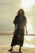 Woman in a black dress standing on a beach with sunlight reflecting off the water.