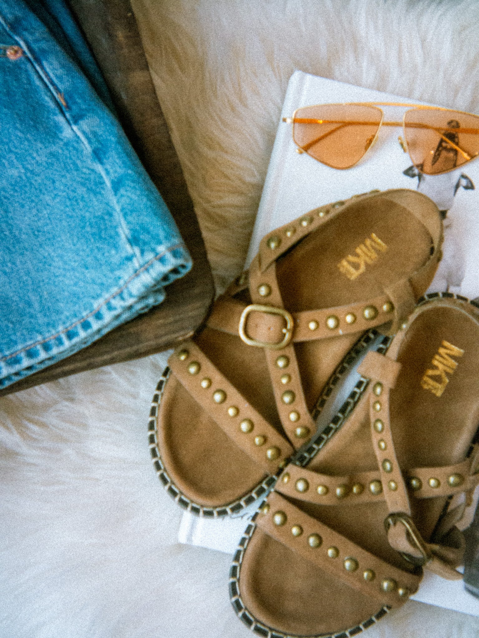 Pair of tan sandals with gold studs on a surface with sunglasses and denim jeans.