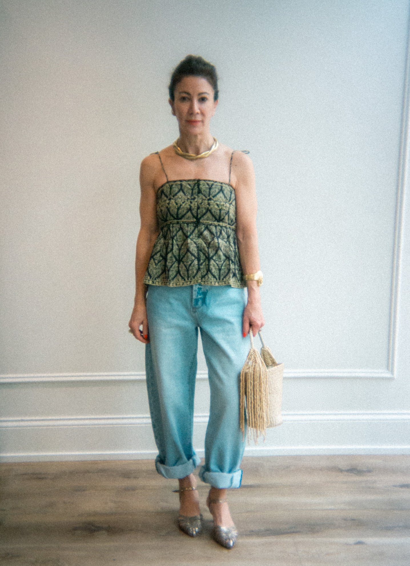 Woman wearing a green patterned top and light blue arc barrel jeans and gold vintage statement necklace, holding a beige bag indoors. 