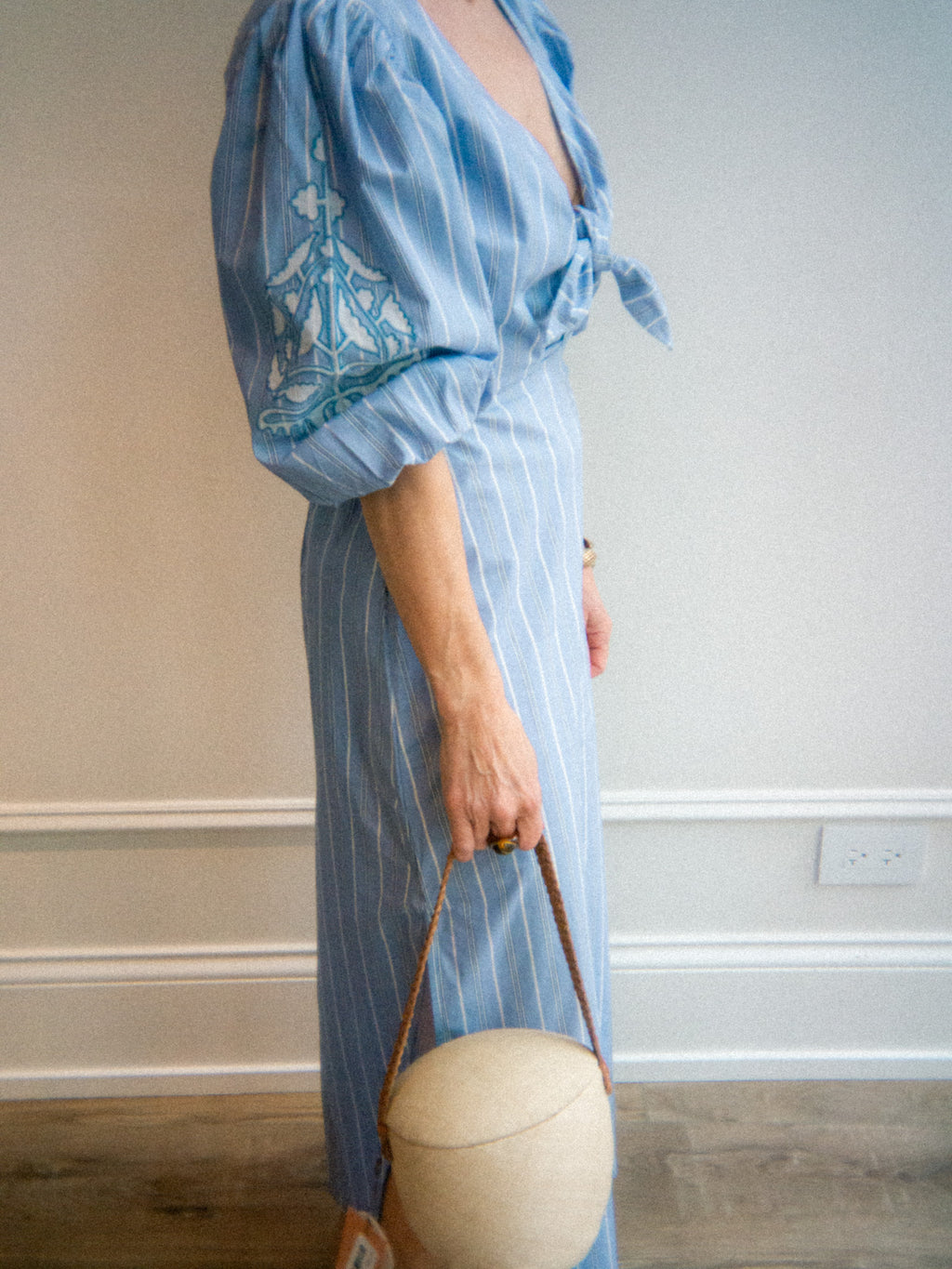 Person wearing a stripe light blue dress with embroidery, holding a beige bag indoors.