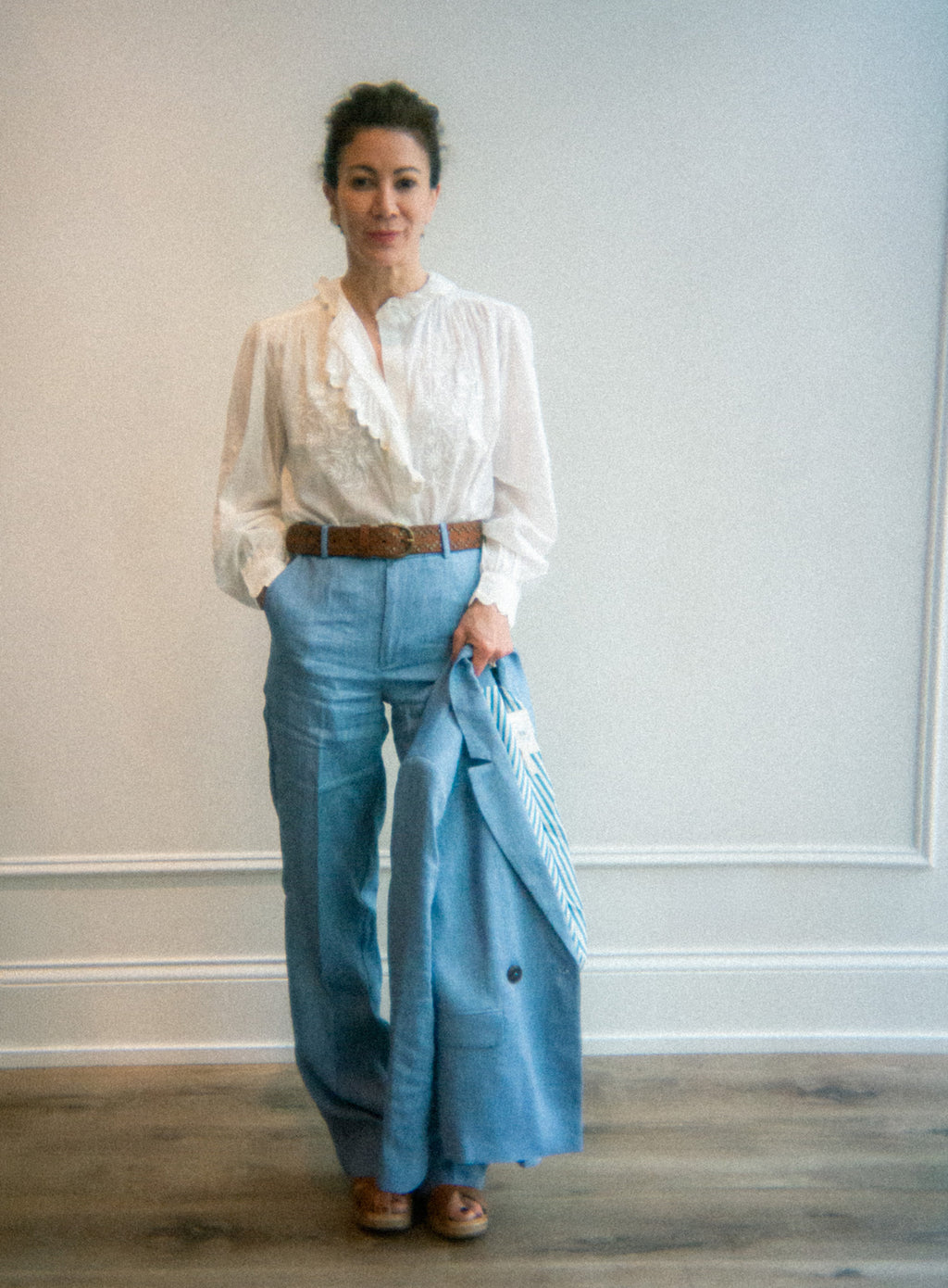Woman wearing a white blouse and blue pants against a plain wall.