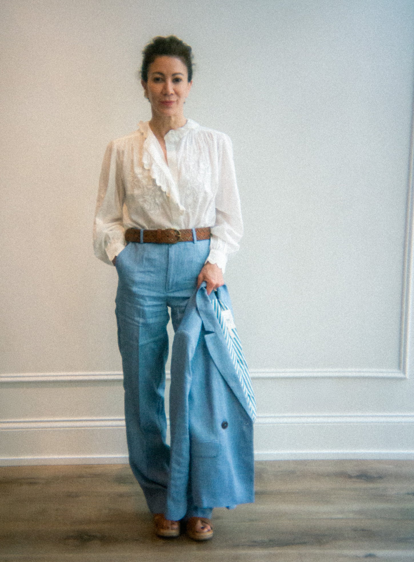 Woman wearing a white blouse and blue pants against a plain wall.
