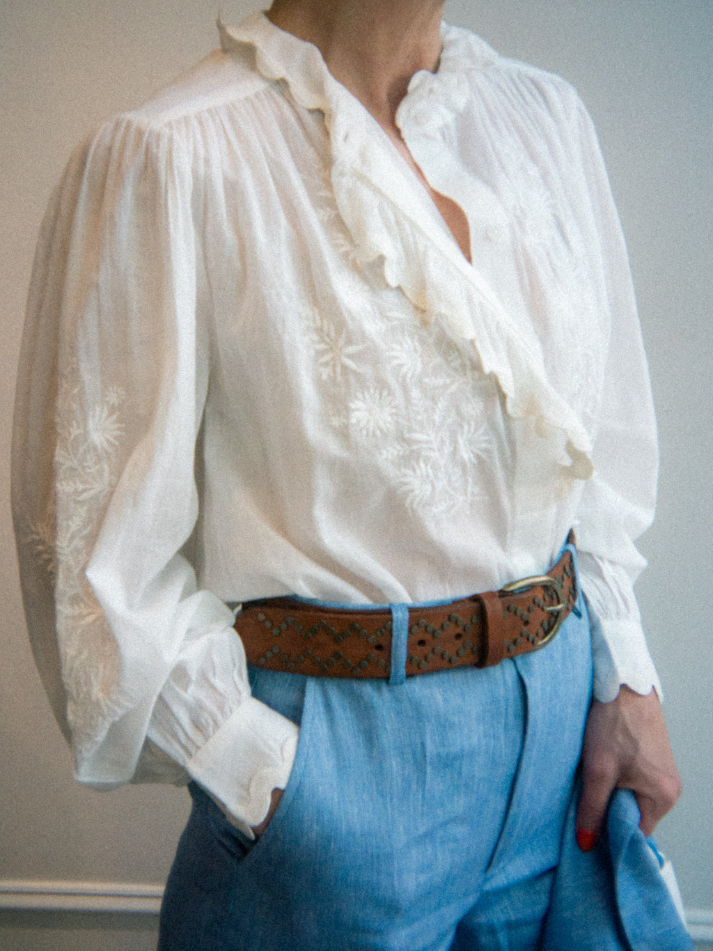 Person wearing a white blouse with ruffled details and blue pants, standing against a plain background.