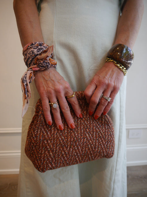 A person in a cream dress holds a brown and orange textured tweed clutch bag. The person is wearing a patterned scarf tied around both wrists and several rings.