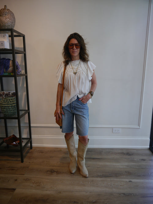 A woman wearing a white short-sleeve blouse, light blue denim shorts, and beige knee-high boots, standing in a room with light wood floors and a white wall.