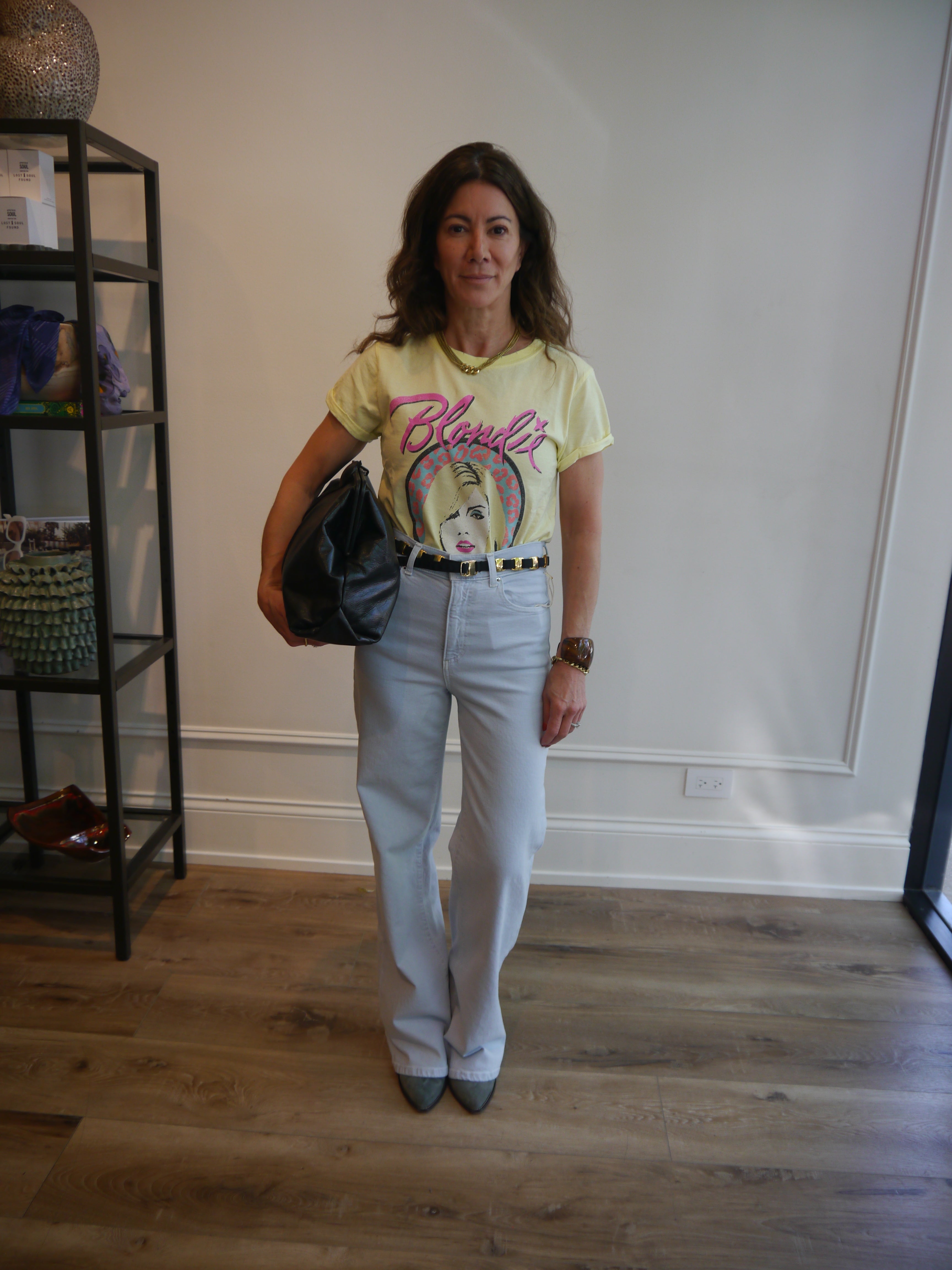 A woman stands indoors wearing a pale yellow graphic T-shirt with pink script lettering, light gray flared jeans, a black belt, and dark pointed shoes. She holds a black handbag and is posed in front of a white wall with wood flooring and a black shelving unit to the left.