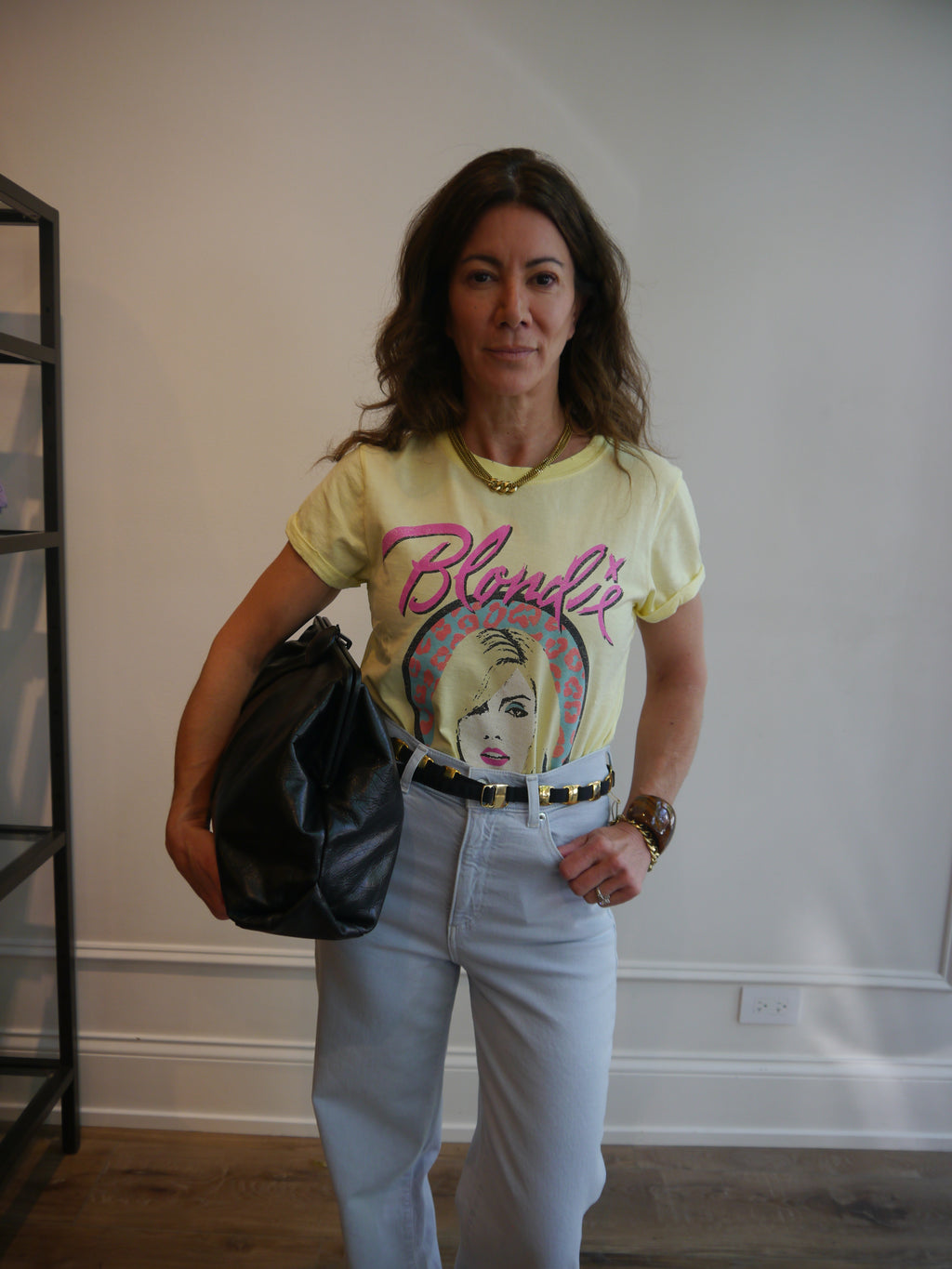 A woman wearing a pale yellow graphic t-shirt with 'Blonde' in pink script and a multicolor portrait print, paired with light blue jeans and holding a black handbag.