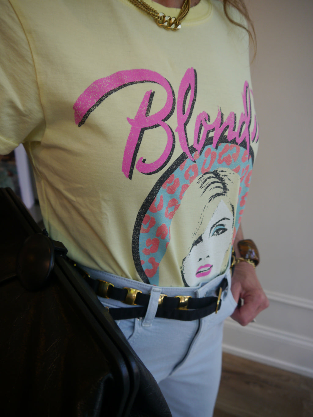 A person wearing a pale yellow graphic T-shirt with a large pink and black 'Blonde' print and a stylized blonde face illustration, paired with light blue jeans, a black belt with gold hardware, and a gold necklace.