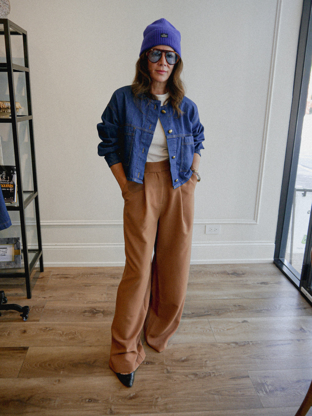 A woman stands in a room wearing a cobalt blue beanie, large vintage oversized sunglasses, a cropped blue denim jacket over a vintage white tee, and high-waisted brown wide-leg trousers.