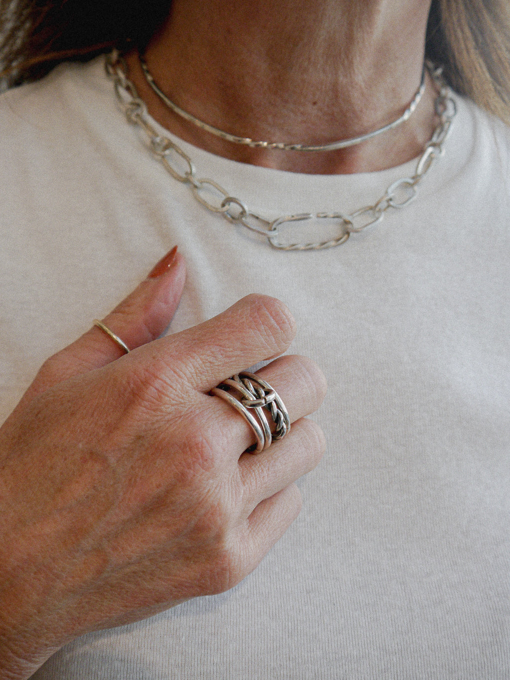 A close-up of a person wearing layered silver jewelry, including a chunky paperclip chain necklace, a thin chain necklace, and multiple silver rings on one finger, over a white t-shirt.