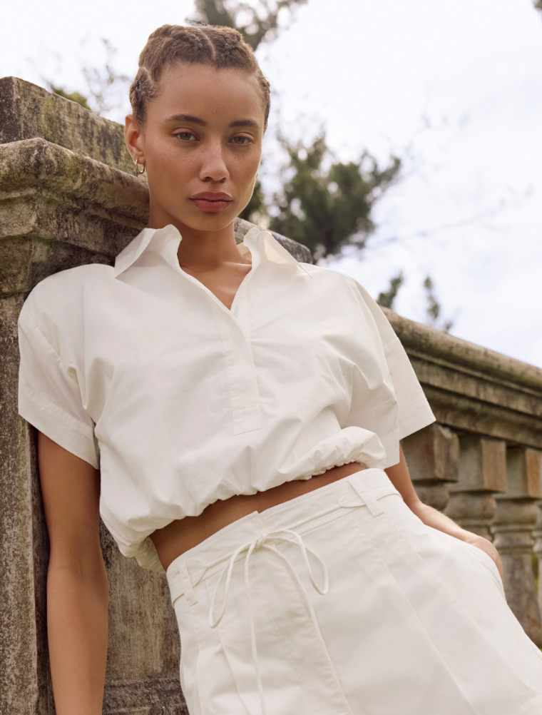 Woman wearing a white outfit leaning against a stone wall outdoors