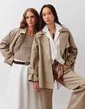 Two women wearing cropped beige trench coats against a white background