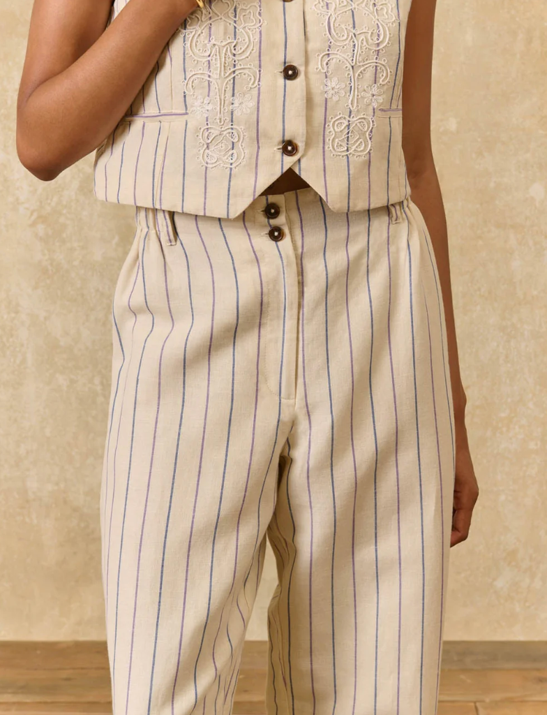 Person wearing a beige vest with intricate embroidery and matching striped pants on a neutral background