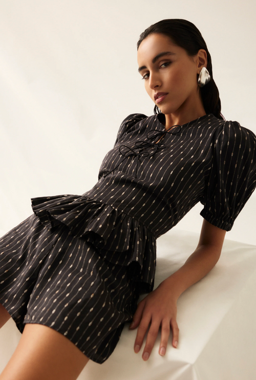 A woman wearing a black short-sleeve dress with a white dotted vertical stripe pattern, ruffled chest detailing, and a peplum waist, posed on a white studio set against a light neutral background.