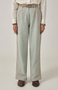 A person wearing light sage green wide-leg trousers with embroidered details on the upper thighs, styled with a white lace blouse, a tan belt, and brown boots against a plain light gray studio background.
