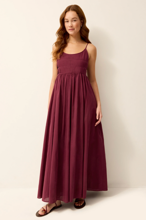 A woman modeling a sleeveless, smocked-bodice maxi dress in deep burgundy with thin spaghetti straps, standing against a plain off-white studio background.