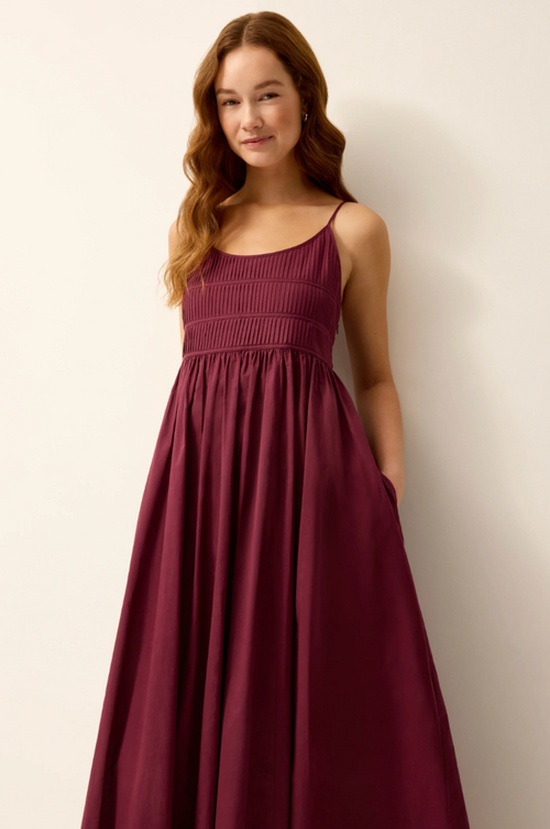 A woman wearing a sleeveless, smocked bodice, floor-length A-line dress in a deep burgundy or maroon color, standing against a plain off-white wall.
