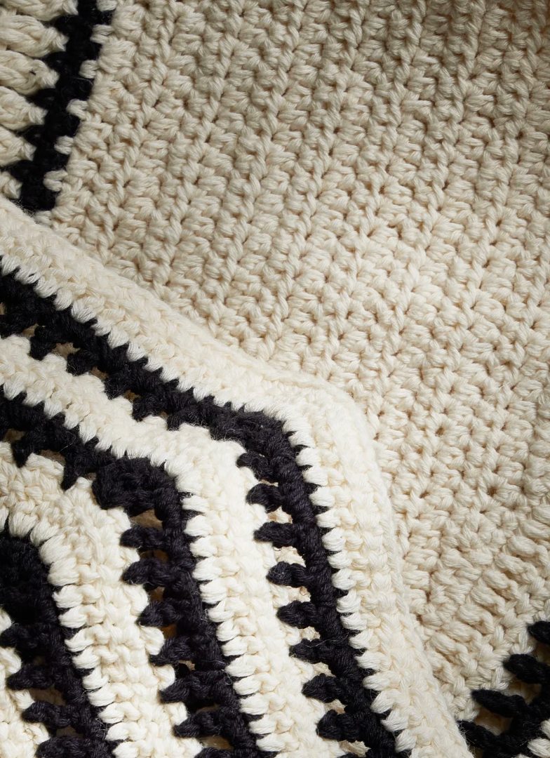 Close-up of a chunky crocheted textile in warm off-white/cream with bold black geometric border patterns.