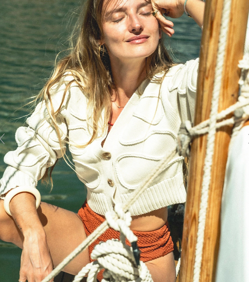 A woman on a boat wearing a cream ribbed knit cardigan and rust-orange shorts, with water and wooden boat rigging in the background.