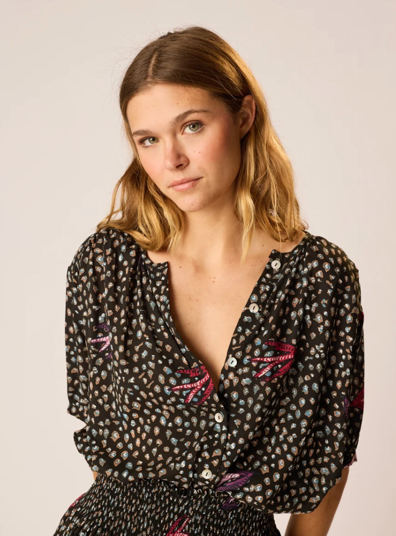 A woman wearing a natalie Martin black short-sleeve blouse with a small multicolor floral print and pink embroidered details, photographed against a plain light background.
