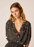 A woman wearing a natalie Martin black short-sleeve blouse with a small multicolor floral print and pink embroidered details, photographed against a plain light background.