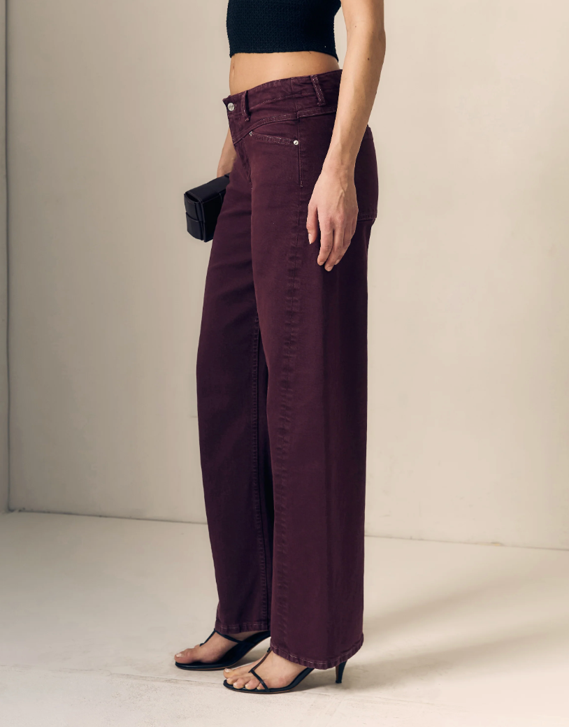 A model wearing a pair of wide-leg, high-waisted pants in a deep plum purple color, styled with a black crop top, black sandals, and holding a small black clutch.