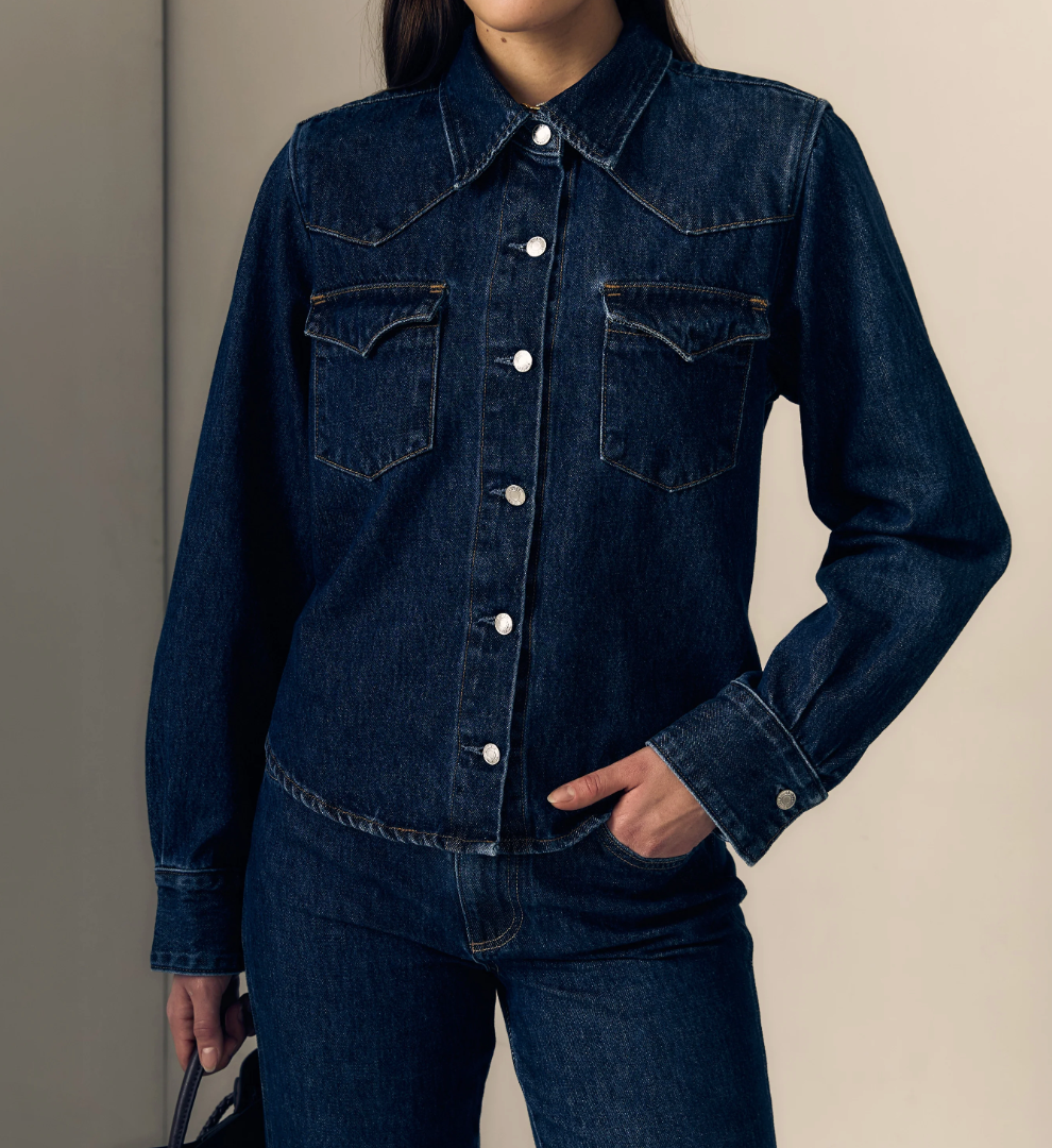 A woman wearing a dark indigo denim long-sleeve button-up shirt with western-style chest pockets and matching dark denim jeans, photographed against a plain beige studio background.