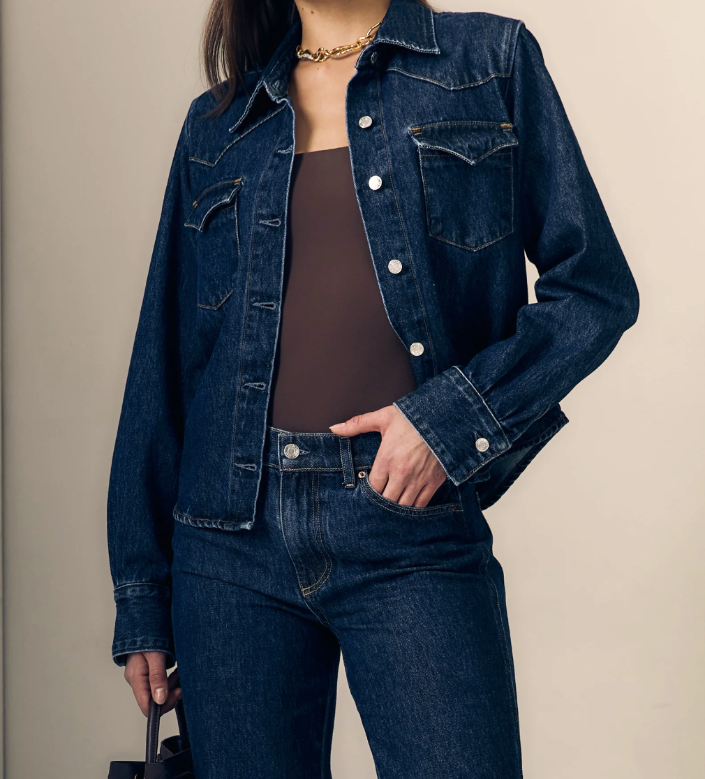 A woman wearing a dark blue denim jacket and matching dark blue jeans over a brown top, standing against a plain beige background.