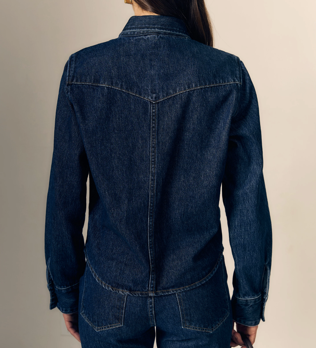 Back view of a person wearing a dark indigo denim jacket and matching denim bottoms against a plain beige studio background.