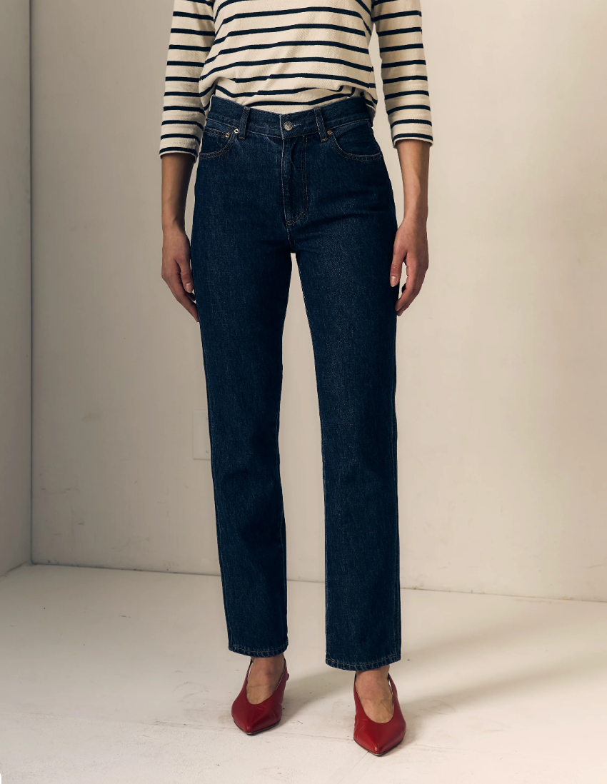 A model wearing dark blue straight-leg denim jeans, a black and white striped long-sleeve top, and red pointed-toe heels, standing against a plain off-white studio background.