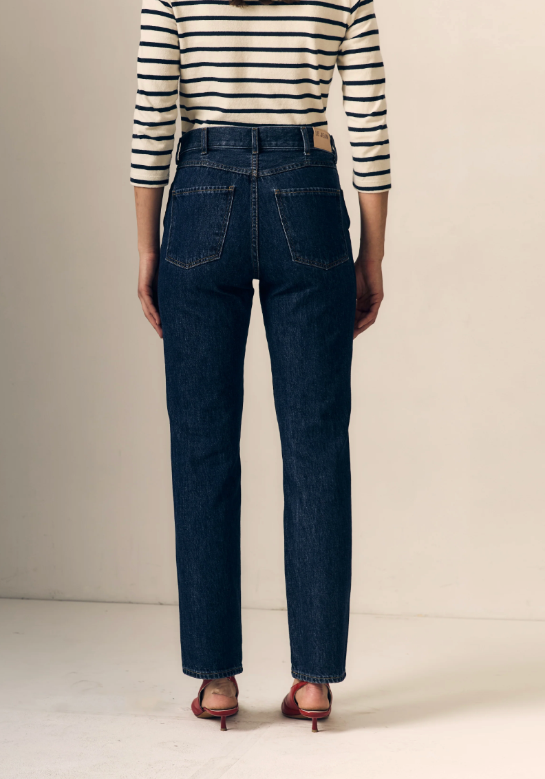 A person shown from the back wearing dark blue straight-leg jeans, a navy and cream striped long-sleeve top, and brown heeled sandals against a plain beige studio background.